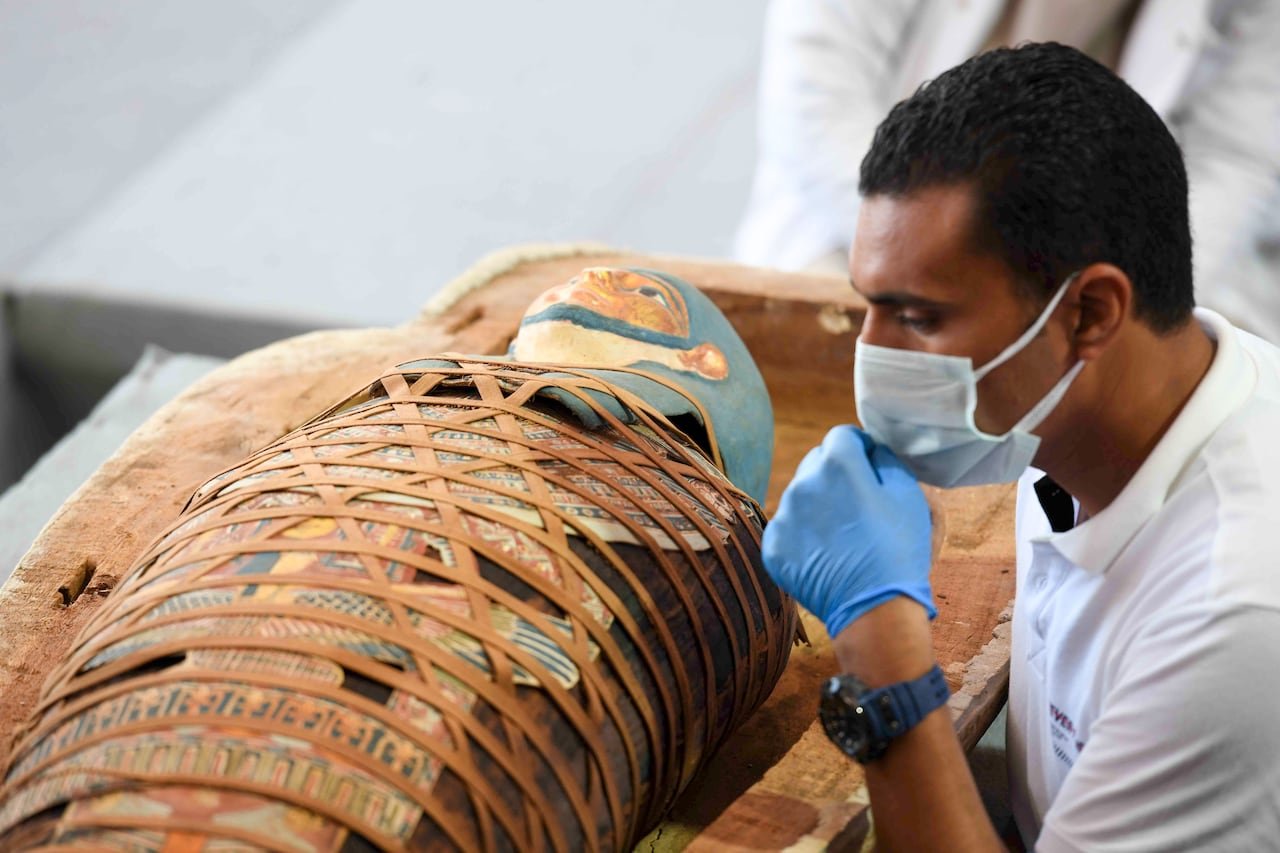 default-973 An archaeologist inspects an Egyptian mummy that is wrapped in an elaborate and colourful burial shroud. The archaeologist is wearing a medical mask and gloves.